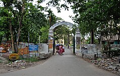 Chemin en asphalte menant à un portail en béton avec écriture en bengali et personnes indiennes en sari marchant à travers le portail.