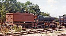 A locomotive is seen from the right and behind. The locomotive is mainly green but there are patches of pale red. On the side of the cab the number 4936 is written in yellow letters. The chimney and boiler dome are missing and the roof of the cabin is detached and is partially resting on the end of the tender. The tender is pale red. Most of the wheels of the locomotive are hidden by a flat wagon. To the right is the end of a tank wagon.