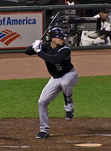 Un uomo con una maglia da baseball nera e pantaloni gessati grigi si prepara per uno swing di baseball con la mano destra.