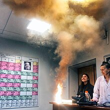 An instructor and a student look at clouds of smoke and a bright flame produced on the edge of a table.