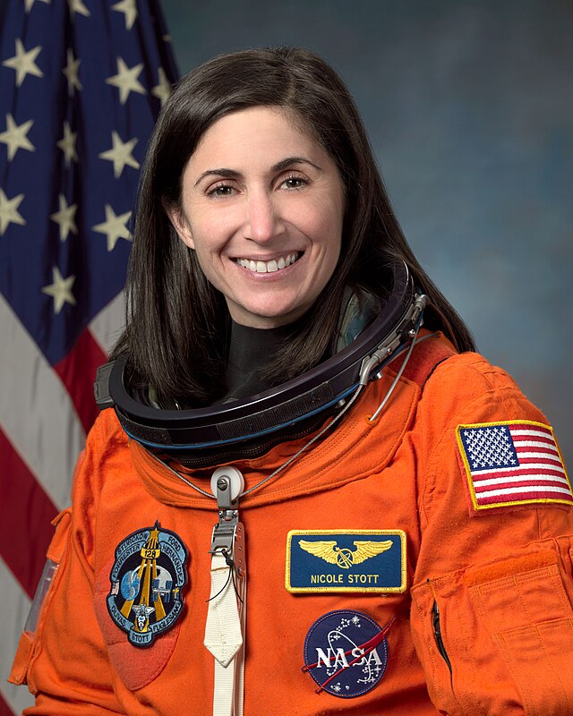 A female Caucasian astronaut wearing her orange mission suit with the American flag embroidered on her shoulder, and name tag and mission patches embroidered on her chest.