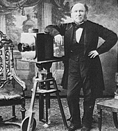 Schwarzweißfotografie eines Mannes, der sich mit seinem rechten Arm an einer Balgenkamera abstützt. Die Kamera steht auf einem Stativ, der ein Rad hat. An beiden Seiten sind Stühle, im Hintergrund eine gemalte Landschaft.
