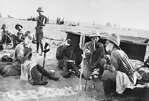 Four men, two wearing pith helmets, sit in the sand, shaving. Three have soap on their faces.