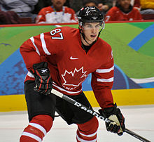 Photographie d'un joueur de hockey sur glace portant un maillot rouge à feuille d'érable avec le numéro 87, casque et short noir et chaussettes rouges, sa crosse en attente de passe.