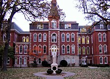 A statue sits in front of a large 4 story red brick building. To the right of the entrance, the building goes in a little ways, and then back out. To the left of the entrance is a small 2 story area connecting the main part of the building to the rest of it. Above and behind the 2 story part is a 4 story part of the building