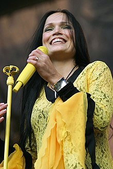 Photo en couleurs d'une femme aux cheveux longs bruns habillée d'un haut en dentelle jaune et noire tenant dans sa main un micro jaune
