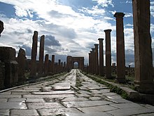 une rue pavée avec des colonnes nues de chaque côté s'étend vers un arc de triomphe au lointain.
