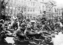 In the foreground soldiers are sitting down with civilians walking behind them. In the distance are four storey buildings