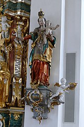 Madonnenstatue mit Krone auf einem Podest vor einer weißen Säule. Auf der gerade nach vorne ausgestreckten Hand der Madonna sitzt das nackte Jesuskind.