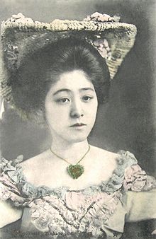 Mladá japonská žena na sobě nabírané šaty a klobouk, s velkým přívěskem nebo medailonem na krku.