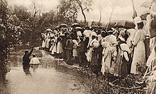 Sepia-toned photograph of a missionary baptising someone in a pond in Brazil, as a crowd watches from the bank.