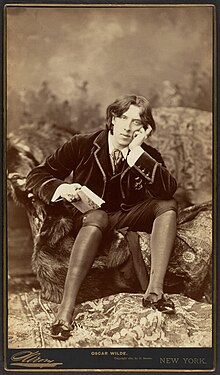 Old photograph of a young man wearing a velvet jacket, knee breeches, silk hose and shiny pointed shoes with bows, seated on a sofa and leaning on his left hand and holding a book in his right