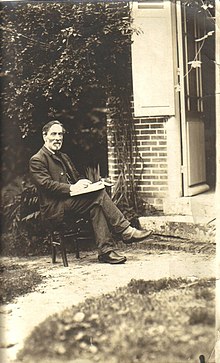 Photographie de Paul Milliet, plus âgé que durant les évènements de la Commune. Il est assis sur une chaise, dehors devant une maison, et tient un livre entre ses mains.