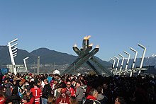 Une foule dense se rassemble devant la vasque olympique. En fond, le ciel bleu et des montagnes.