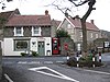 The markings on a road junction around a tree are visible in the foreground, in front of a small shop with Christmas decorations in the window. A red K8 model telephone box and a red post box built into a wall are to the left.