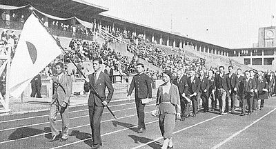 Photo noir et blanc d'une délégation sportive défilant dans un stade. Au premier plan, un porte-drapeau tenant l'emblème national japonais.