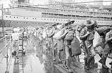 Men in shorts and slouch hats with rifles slung, carrying duffel bags march along a wharf. In the background is a cruise ship.