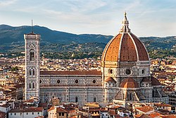 The Cathedral of Saint Mary of the Flower, illuminated at night, showing the large red brick dome, a decorated white marble nave, and a vertical, white marble bell tower to the left. Mountains are visible in the background and a dark, low-lying city in the foreground.