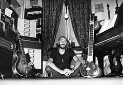 Un homme barbu aux cheveux longs assis par terre dans un appartement rempli d'instruments de musique et d'appareils d'enregistrement.