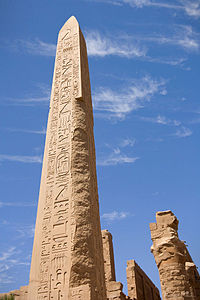 Pískovcový obelisk pokrytý hieroglyfy. V pozadí se nacházejí pískovcové trosky chrámu.