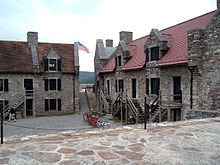 A photograph of two stone buildings with red roofs, surrounding a stone-paved central area. The buildings have entrances on two levels, with wooden stairs outside leading to the doorways on the upper level. An American flag is visible waving in the gap between the buildings.