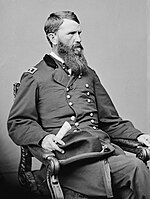 Black and white photo of a bearded man seated in a chair and holding a rolled up piece of paper in his right hand. He wears a dark military uniform with two rows of buttons.