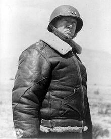 Portrait en contre-plongée de Patton portant un casque avec trois étoiles sur le devant et une veste de cuir rembourrée.