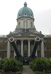 A three-storey brick building with tall copper dome on top, a frontage of six Doric columns and portico, and in front of the columns two large naval guns with tampion badges on a concrete base.