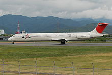 A McDonnell Douglas MD-81 aircraft taxiing on the taxiway, with grass field and a grey coloured barbed wired fence in the foregound, and a mountain view on the background