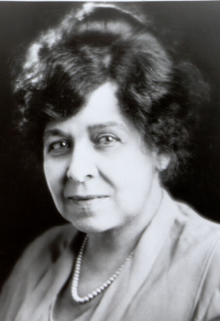 A portrait of a Black woman with light skin, hair dressed in an updo, wearing a light-colored, soft blouse with a strand of pearls