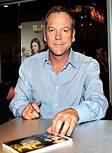 A colored photograph of a blonde-haired man with a blue shirt. He is looking directly at the camera, with a pen on his right hand and book visible on the table.