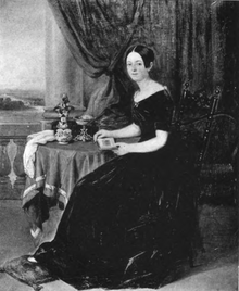 Full-length portrait of Louise Milliet, mother, in dress, seated in an armchair with her feet on a cushion on the floor. She is leaning on a table and holding a notebook in her hands. Several objects are arranged on the table.