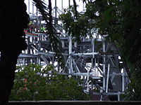 Structure métallique visible à travers des feuillages, avec deux ouvriers portant des gilets jaunes.