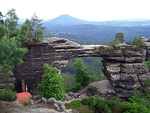 Na pierwszym planie skalne płyty tworzą most, pod którym przestrzeń jak otwarta brama. Skały są otoczone zielenią. U dołu zdjęcia widoczny fragment czerwonego dachu, ponad skałami w oddali widoczny szczyt górski.