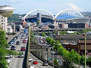 An elevated highway with inbound traffic seen atop a double-decked structure. Several buildings can be seen off to the side of the highway, while CenturyLink Field and Mount Rainier can be seen in the background.