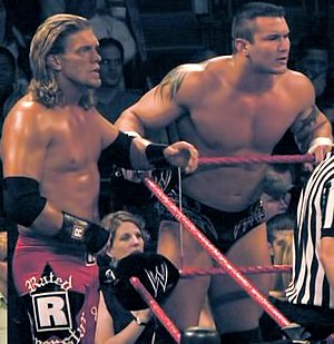 Two Caucasian males stand on the corner of a wrestling ring apron with red ropes. The one closest to the camera has shoulder-length blond hair and is wearing long black wrestling tights with the words "Rated R Superstar" detailed upon them in white. The other has short dark hair, is wearing short black wrestling tights, and tattoos are visible on his upper arms.