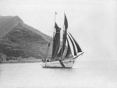 Photo noir et blanc d’un bateau qui navigue à la voile près d’une côte abrupte.