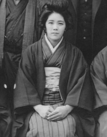 A black and white photo of a young woman, sitting, wearing a striped kimono and a jacket and a traditional hairstyle.