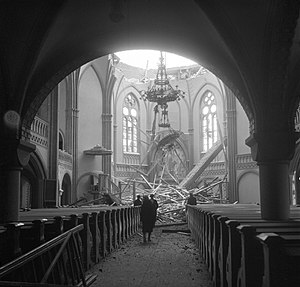 The dome of the Viipuri Cathedral collapsed after the Soviet bombing. Four people stand in the nave and look at the rubble, which is highlighted by sunlight shining through the damaged dome.