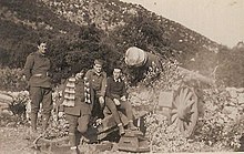 Vier Soldaten sitzen vor einer großen Kanone mit großen Rädern in einer bergigen Landschaft.