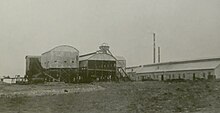 Photographie en noir et blanc de plusieurs hangars et bâtiments couverts arborant des cheminées, et ce qui semble être l'accès au puits de remontée vers la surface.