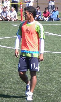 A man wearing a green t-shirt, orange bib and navy blue shorts, standing on a soccer field. In the background, people are sat watching him.