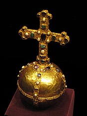 A smooth gold sphere wrapped in raised bands along the equator and two perpendicular longitudinal rings. A cross, taller than the orb's diameter is mounted on top. The bands and cross are garishly embossed with a curly vine pattern and studded with gems.