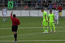 Two players stand over a soccer ball at the center of the field while a referee holds his hand over head.