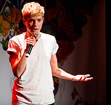 Junge, blonde Person in weißem T-Shirt mit Mikrofon auf kleiner Bühne in einem Club