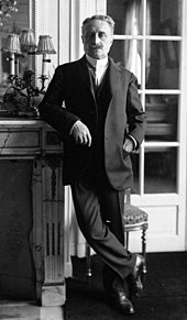 Photo en noir et blanc d'un homme se tenant debout, accoudé à une cheminée, la main gauche dans la poche de sa veste ; moustache et cheveux grisonnants, chevilles croisées, il porte un costume trois pièces, une chemise blanche à faux col et une cravate
