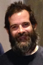 En middelaldrende mann med brunt hår og et busket brunt skjegg som smiler til kameraet.