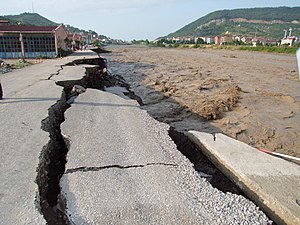 road with large cracks being washed away by a roiling brown river