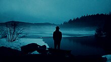 silhouette of a man wearing an overcoat and fedora before foggy river at dusk.