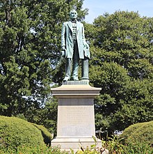 A color photograph of a greened bronze statue atop a granite pedestal